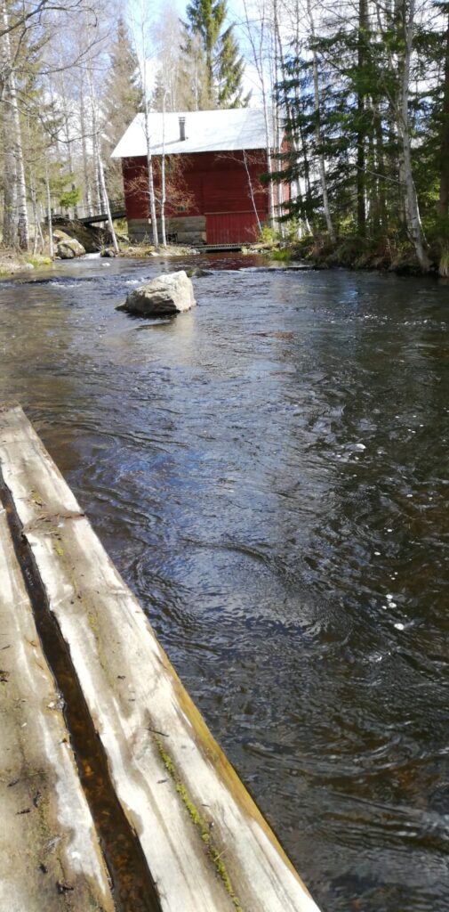 Mylly alajuoksulta päin. Näkyy pieni osa melontalaiturin lautoja ja iso kivi keskellä virtaa.